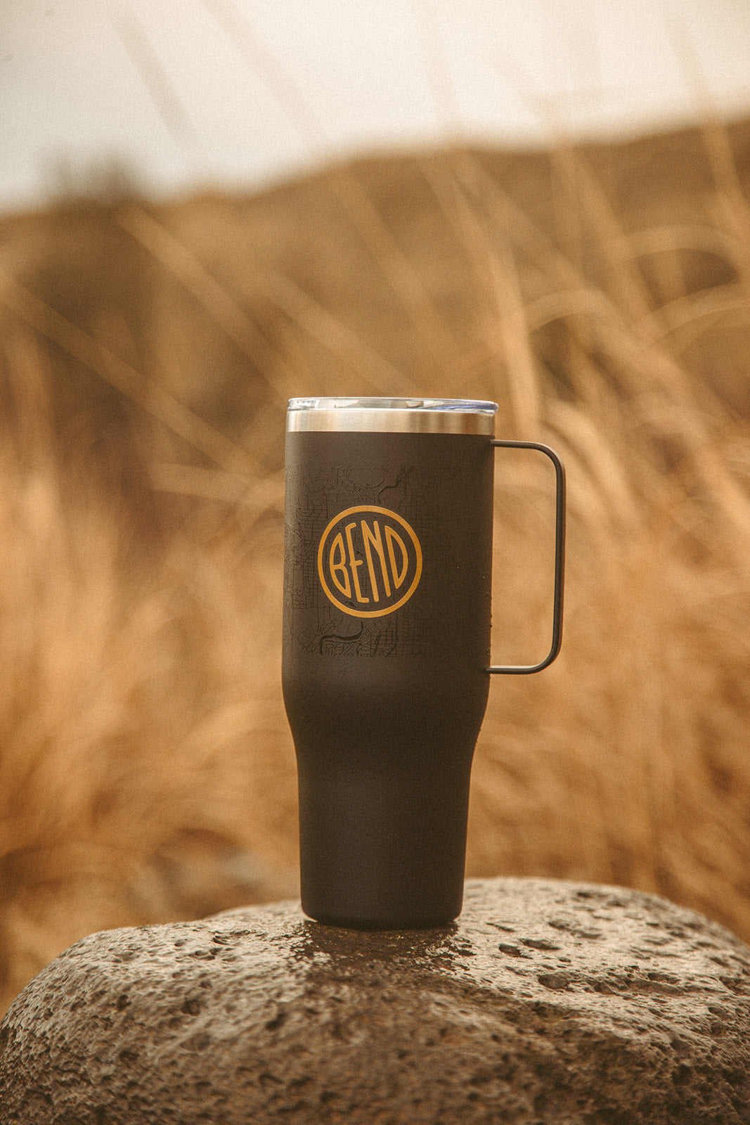 Black insulated mug with a logo on a rock against a blurred natural background