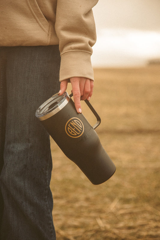 Person holding a black insulated mug with a logo of Bend Oregon in a field
